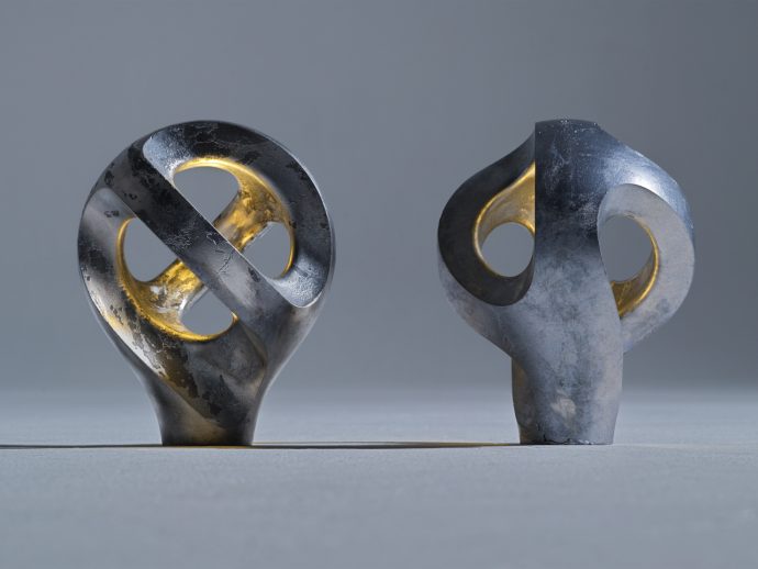 Two weathered metal sculptures with circular cutouts stand on a gray surface in a neutral studio, gold inner rings visible through the holes.