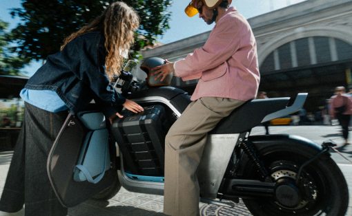 Two people with a light-colored scooter: one in a pink jacket sits on the bike while the other helps open the side panel.