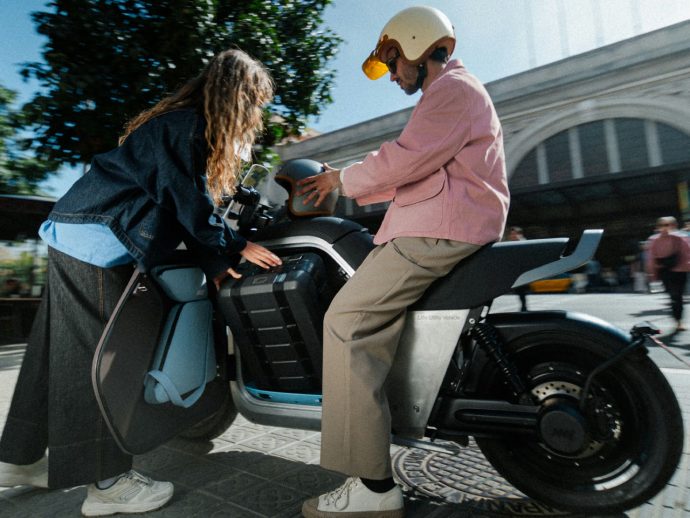 Two people with a light-colored scooter: one in a pink jacket sits on the bike while the other helps open the side panel.