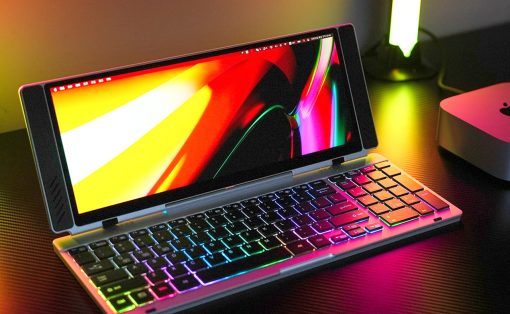 Compact laptop in tent/stand mode with a vivid rainbow RGB keyboard backlight on a dark desk surface, screen displaying bright colors.