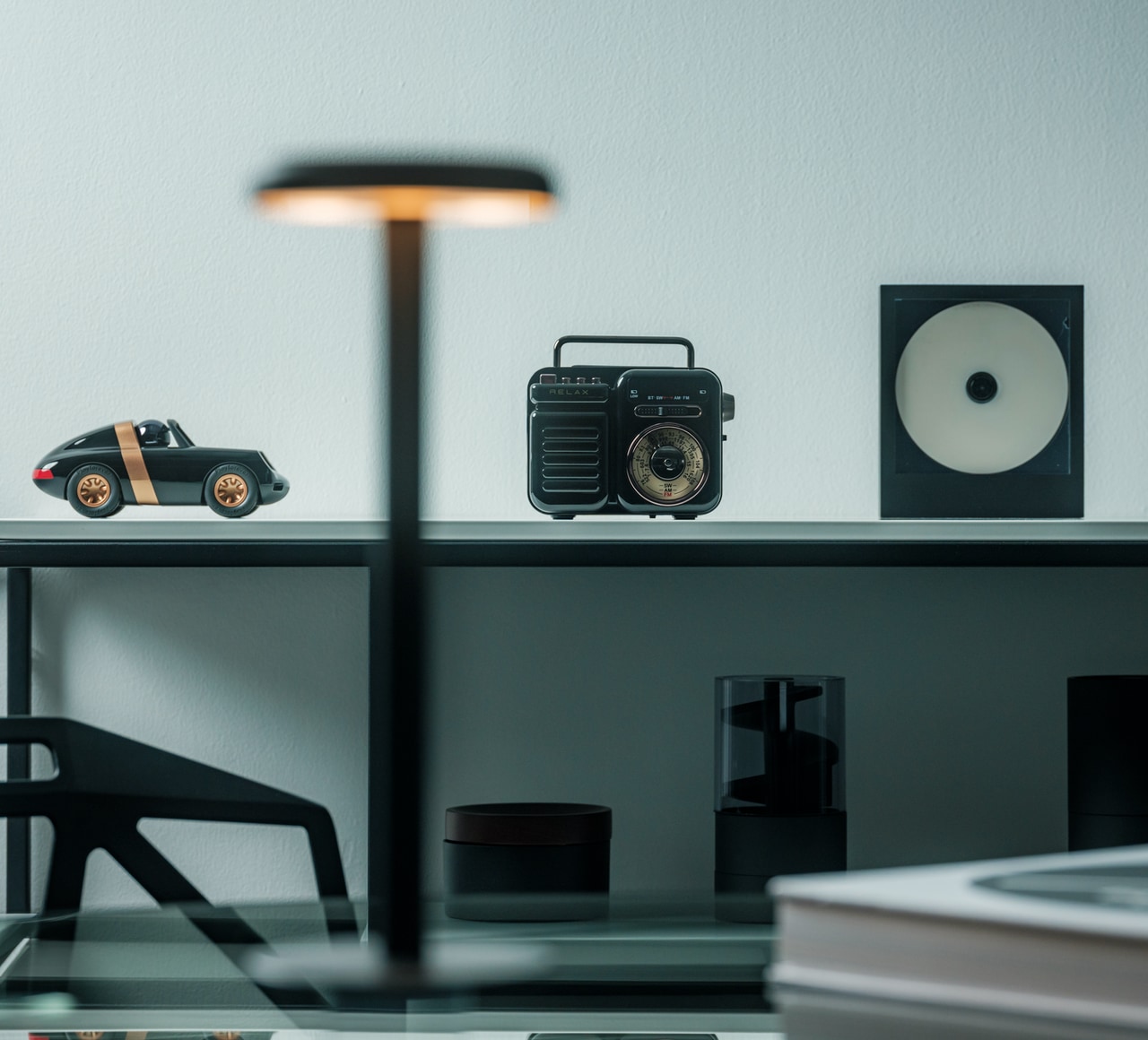 A vintage portable radio sits on a shelf between a black toy car and a square speaker with a white disc on the right of the image.