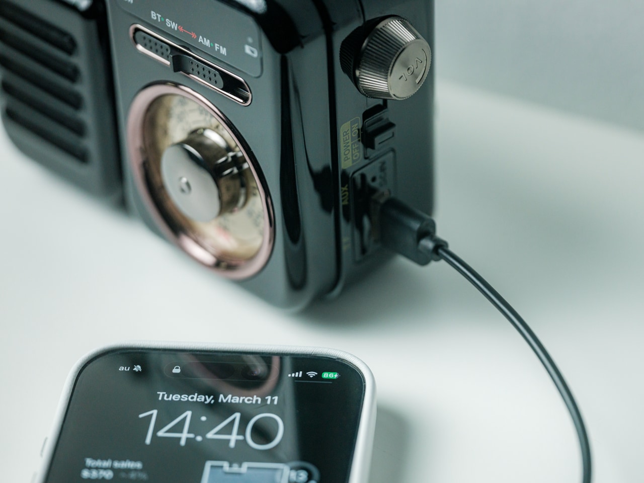 Close-up of a vintage black radio with a charging cable plugged into a smartphone displaying 14:40 on its screen.