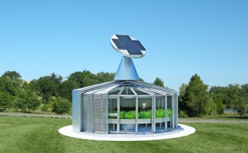 Circular glass pavilion with a solar-powered turbine on a blue cone, set on a white circular base in a grassy park