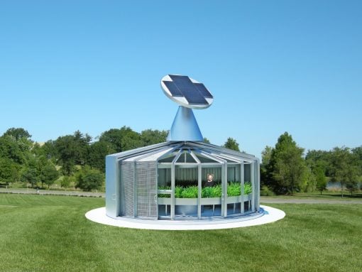 Circular glass pavilion with a solar-powered turbine on a blue cone, set on a white circular base in a grassy park