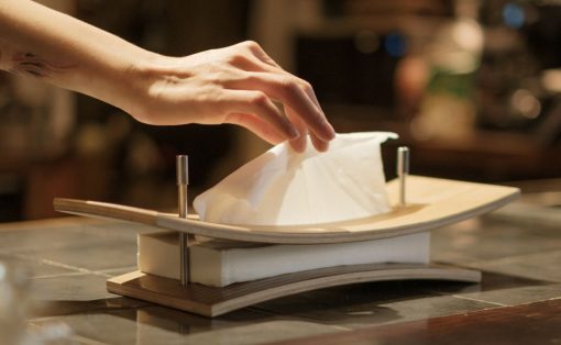 A hand places a white napkin on a curved wooden plate set on a napkin holder atop a marble counter in a dim restaurant.