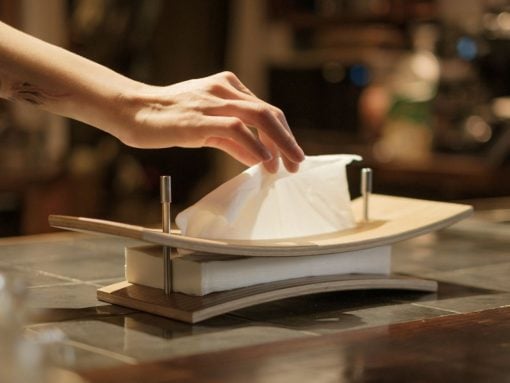 A hand places a white napkin on a curved wooden plate set on a napkin holder atop a marble counter in a dim restaurant.