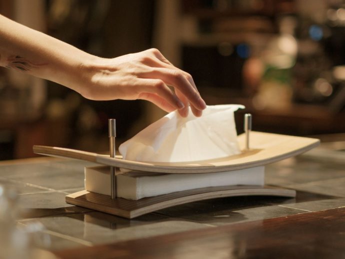 A hand places a white napkin on a curved wooden plate set on a napkin holder atop a marble counter in a dim restaurant.