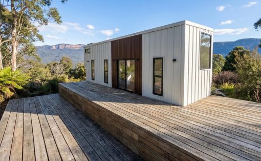 Modern white tiny house on a multi-level wooden deck with a mountain landscape in the background.