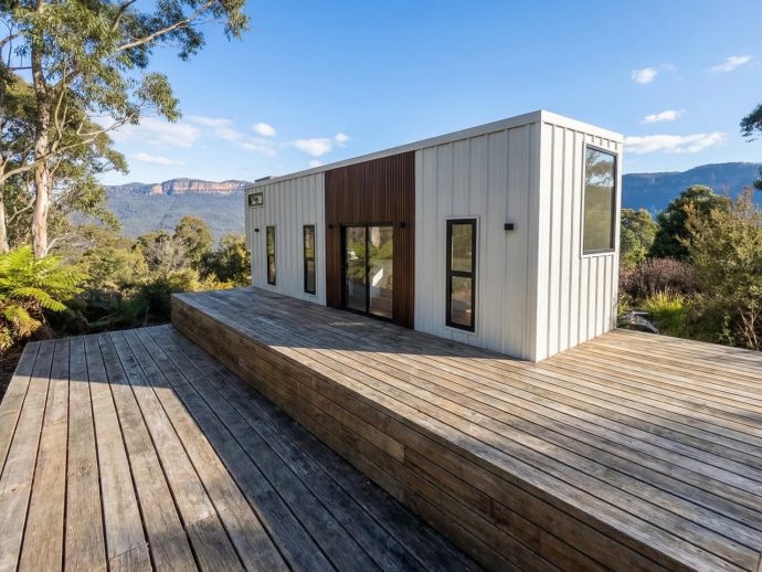 Modern white tiny house on a multi-level wooden deck with a mountain landscape in the background.