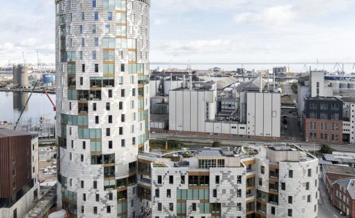 Tall cylindrical modern building with a glass and metal facade beside a harbor and industrial district.