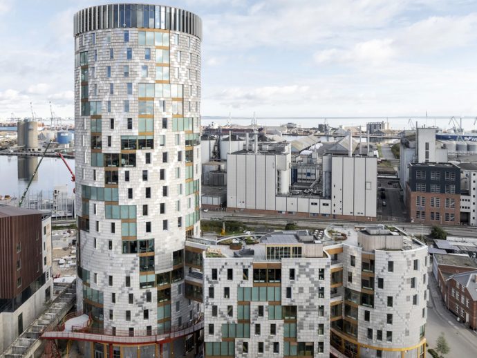 Tall cylindrical modern building with a glass and metal facade beside a harbor and industrial district.