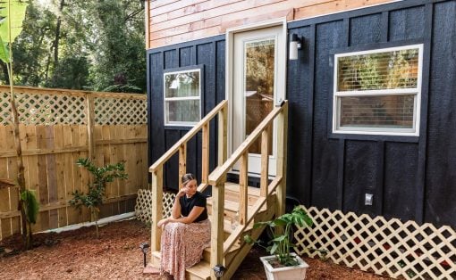 Woman sits on wooden steps of a tiny house with black siding and orange trim in a fenced, mulched yard