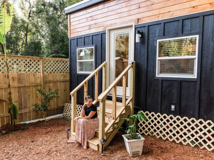 Woman sits on wooden steps of a tiny house with black siding and orange trim in a fenced, mulched yard