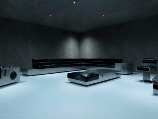 Dark minimalist living room with a black sectional sofa, low square ottoman, and sleek metal accents on a bright pale floor.