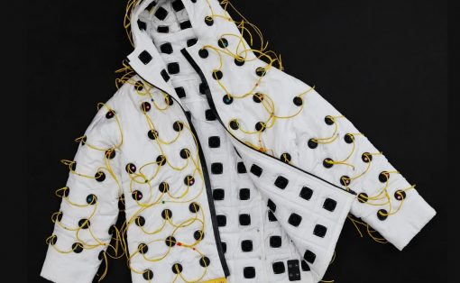 White quilted jacket covered in numerous black square panels connected by yellow wires, laid against a dark backdrop, with a yellow tag reading 'Vollebak Sonic Jacket' nearby.