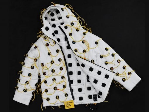 White quilted jacket covered in numerous black square panels connected by yellow wires, laid against a dark backdrop, with a yellow tag reading 'Vollebak Sonic Jacket' nearby.