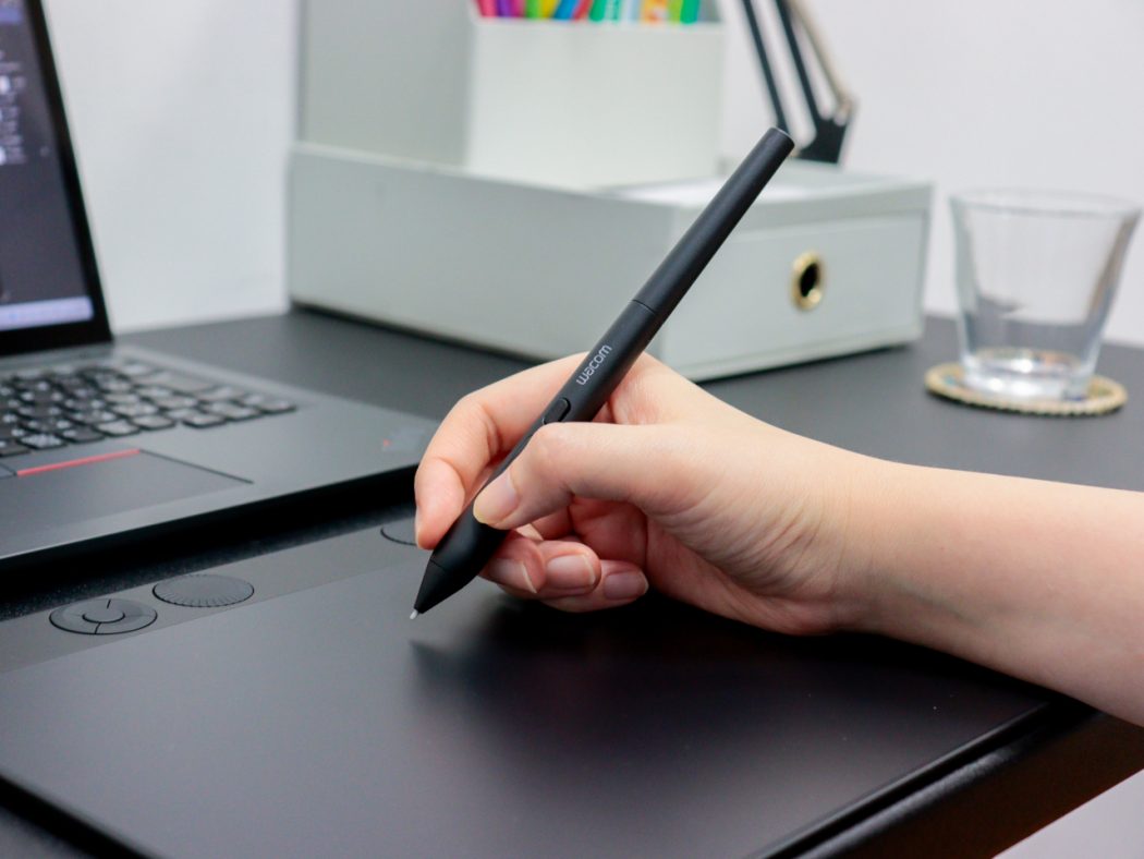 Hand holding a Wacom stylus over a graphic tablet on a desk beside a laptop and office items