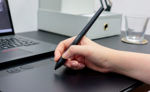 Hand holding a Wacom stylus over a graphic tablet on a desk beside a laptop and office items