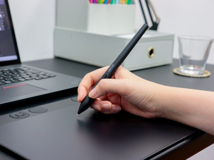 Hand holding a Wacom stylus over a graphic tablet on a desk beside a laptop and office items