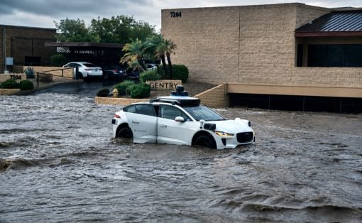White Waymo self-driving car partially submerged in floodwater on a street in front of a beige brick building with palm trees and parked cars nearby; floodwaters cover the road and surrounding area, suggesting severe rain or storm impact.