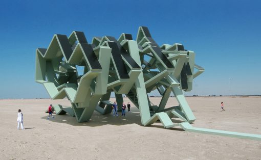 Large pale green angular metal sculpture in a barren desert, with several people for scale.