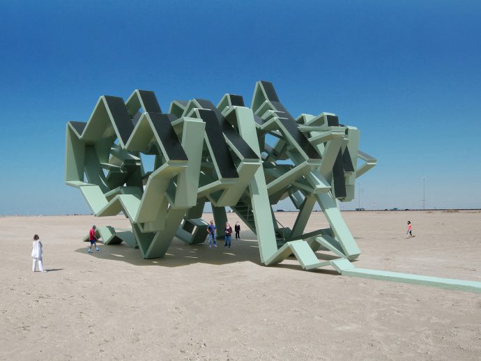 Large pale green angular metal sculpture in a barren desert, with several people for scale.