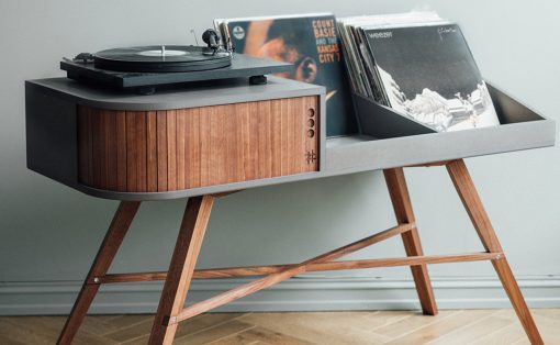 Mid-century wooden turntable on a gray console with a stack of vinyl records in a tray behind it.