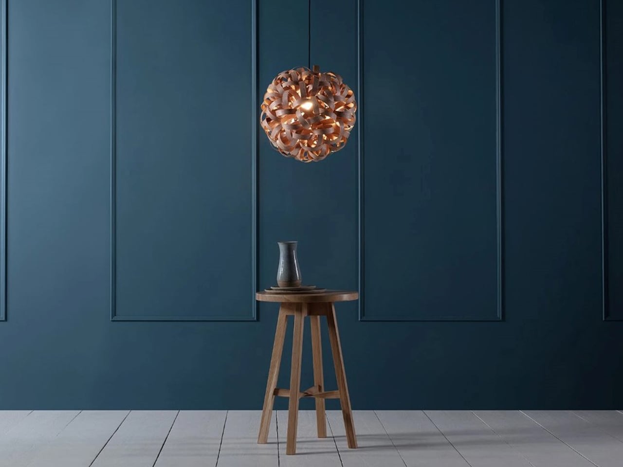 Wooden stool with a small ceramic vase under a copper pendant light against a dark blue paneled wall.