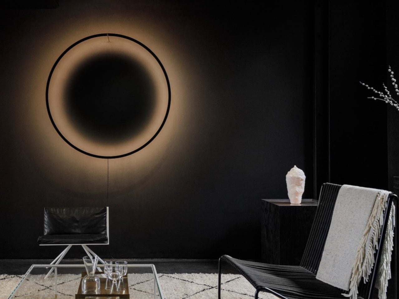 Dark minimalist living room with a large circular wall halo, a black chair, and a glass coffee table; a white sculpture rests on a side table to the right.