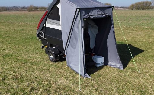 Compact camping trailer with a gray tent annex set up on a grassy field under a clear blue sky