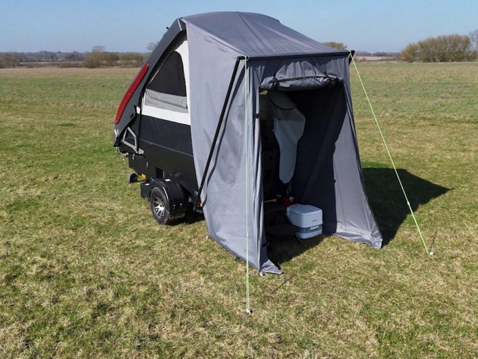 Compact camping trailer with a gray tent annex set up on a grassy field under a clear blue sky