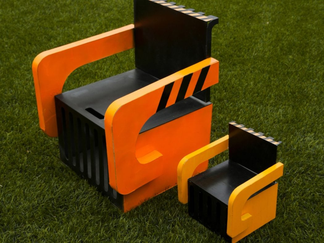 Abstract outdoor sculpture of stacked orange and black blocks with curved orange rails on a grassy field.