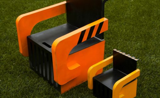 Abstract outdoor sculpture of stacked orange and black blocks with curved orange rails on a grassy field.