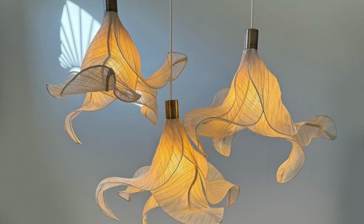 Three sculptural pendant lamps with twisting translucent fabric shades emitting warm light. Two taller and one shorter, suspended from cords against a pale wall.