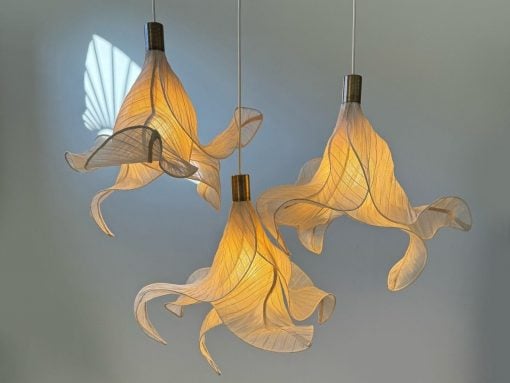 Three sculptural pendant lamps with twisting translucent fabric shades emitting warm light. Two taller and one shorter, suspended from cords against a pale wall.