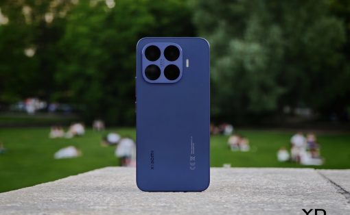 Blue Xiaomi smartphone with a four‑camera square module standing upright on a stone ledge in a park.