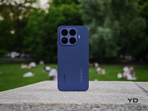 Blue Xiaomi smartphone with a four‑camera square module standing upright on a stone ledge in a park.