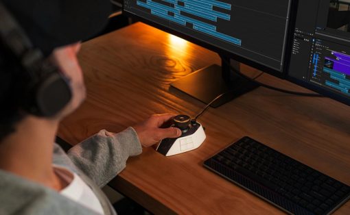 Person with headphones using a compact editing controller at a desk with two monitors showing video timelines.