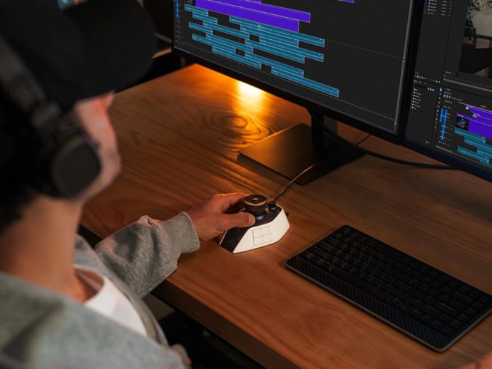 Person with headphones using a compact editing controller at a desk with two monitors showing video timelines.