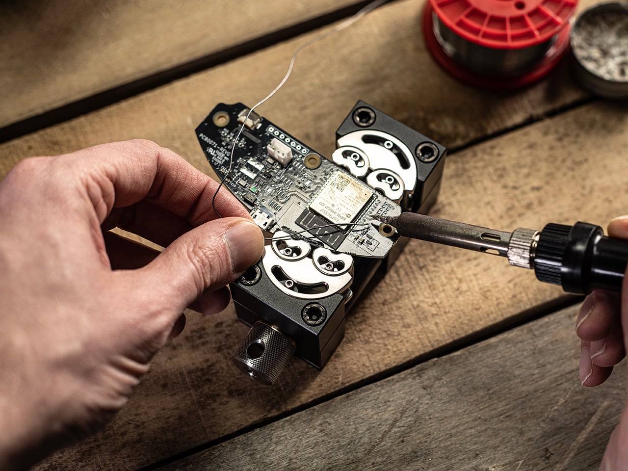 Person soldering a small circuit board secured in a vise on a wooden workbench, soldering iron touching a component.