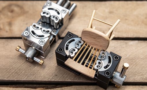 Close-up of a metal precision clamp/jig with screws on a wooden work surface, a small wooden chair-like piece resting on it.