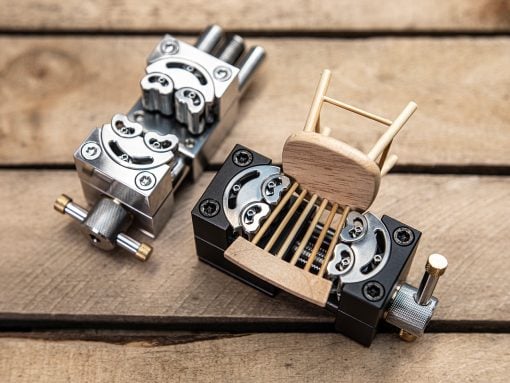 Close-up of a metal precision clamp/jig with screws on a wooden work surface, a small wooden chair-like piece resting on it.