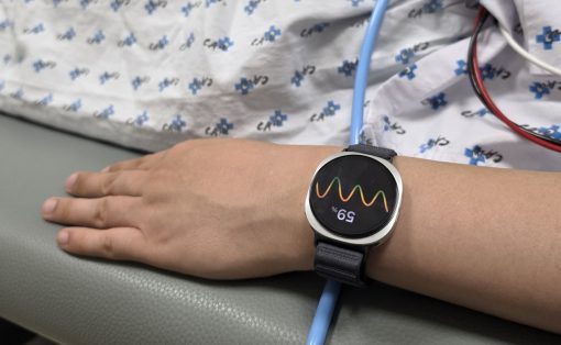 Wrist wearing a round smartwatch showing a colorful wave pattern on a hospital bed, with patient in patterned gown nearby and medical tubing.
