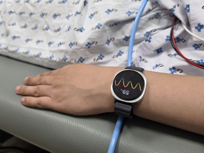 Wrist wearing a round smartwatch showing a colorful wave pattern on a hospital bed, with patient in patterned gown nearby and medical tubing.