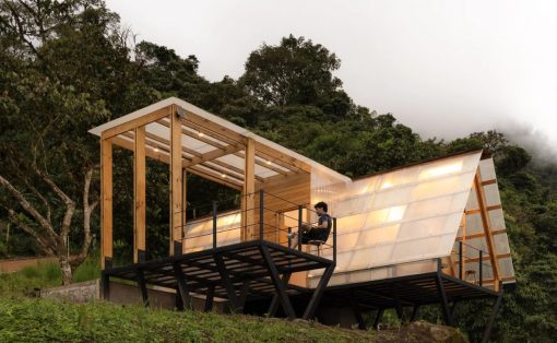 Modern wooden cabin on stilts with glass walls on a hillside; a person sits on the deck chair.