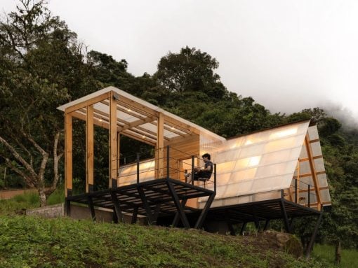 Modern wooden cabin on stilts with glass walls on a hillside; a person sits on the deck chair.