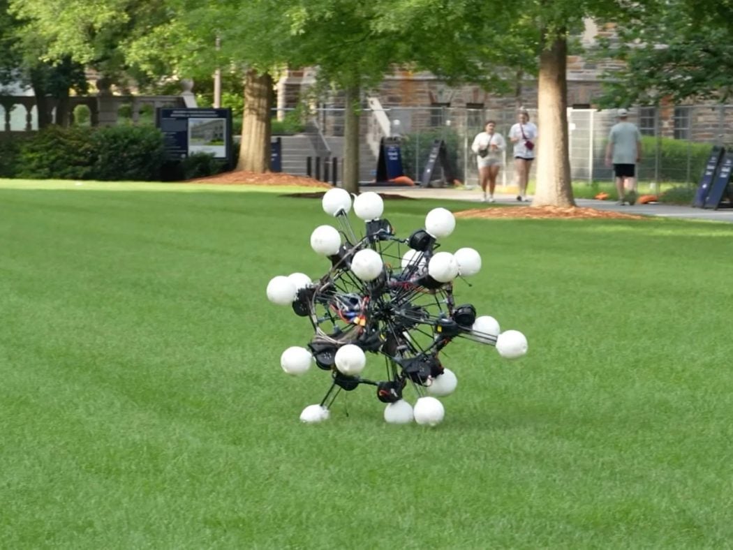Experimental spherical drone with multiple white pods on a grassy university lawn, with people walking in the background.