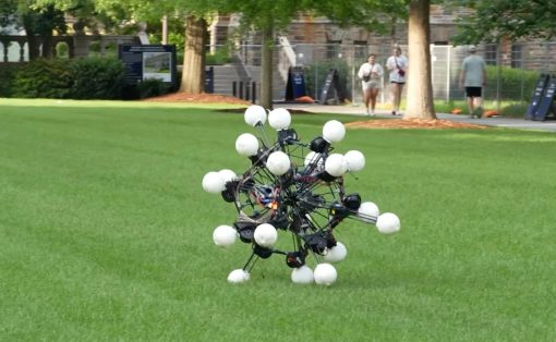 Experimental spherical drone with multiple white pods on a grassy university lawn, with people walking in the background.