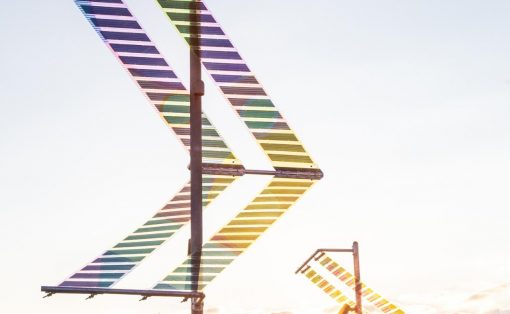 Rainbow-striped metal sculpture forming a large chevron against a pale sky, outdoor art installation.