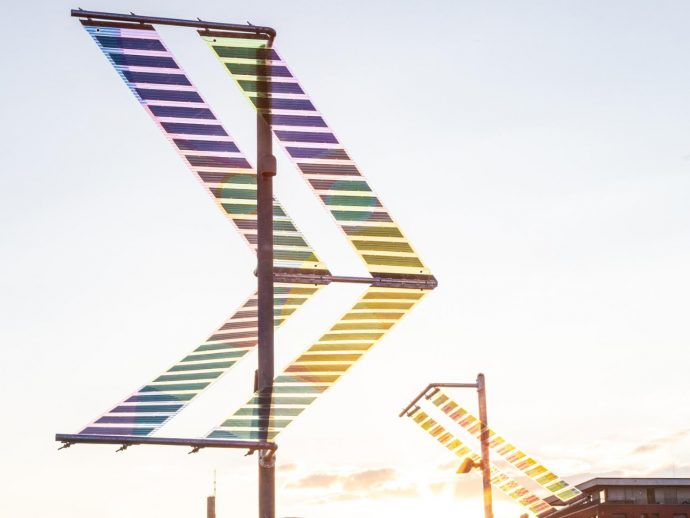 Rainbow-striped metal sculpture forming a large chevron against a pale sky, outdoor art installation.